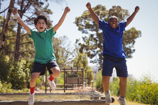 Two boys are cheering and jumping on an obstacle course in a boot camp. They are outdoors, surrounded by trees and greenery, enjoying a sunny day. This image can be used for promoting outdoor activities, summer camps, teamwork, children's fitness programs, and adventure sports.