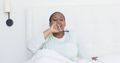 Yawning african american woman waking up in minimalist white bedroom wearing soft pajamas