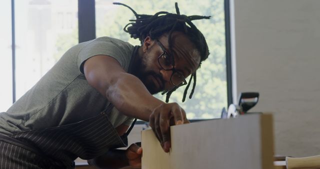 Man with glasses and dreadlocks carefully measuring a piece of wood in well-lit workshop. Ideal for themes around craftsmanship, woodworking, DIY projects, artisan skills, handmade goods, and precision labor. Can be used in articles, advertisements, or instructional content related to carpentry and craftsmanship.