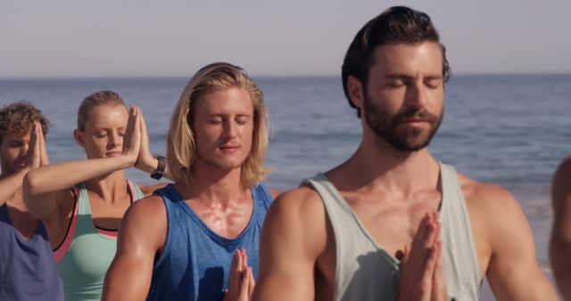 Group of adults practicing yoga and meditation on the beach near the ocean, wearing casual athletic wear. Useful for content related to wellness, outdoor activities, group fitness, mindfulness practices, and promoting a healthy lifestyle.