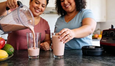 Two women pouring fruit smoothies on kitchen counter, smiling and sharing healthy morning