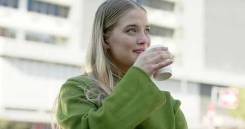 Young woman savoring takeaway coffee on urban street wearing green coat