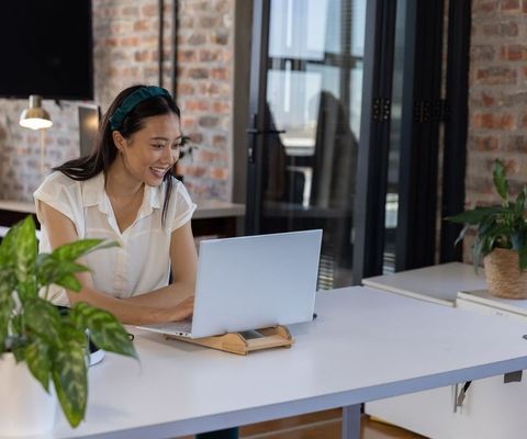 Asian businesswoman smiling while working on laptop at modern bright office desk