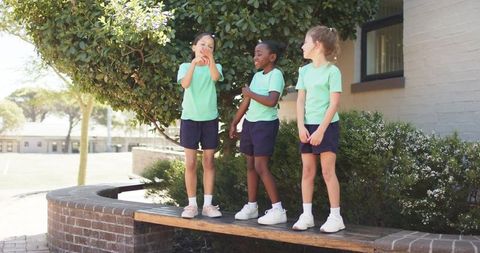 Multicultural girls standing on bench in sunlit school courtyard wearing green shirts and navy short
