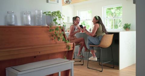 Two women laughing over phone at marble kitchen island with wooden piano and green plants