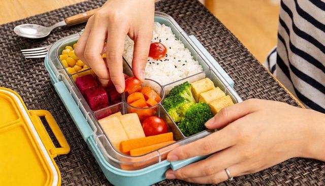 Woman picking cherry tomato from colorful bento lunchbox with rice, broccoli, tofu
