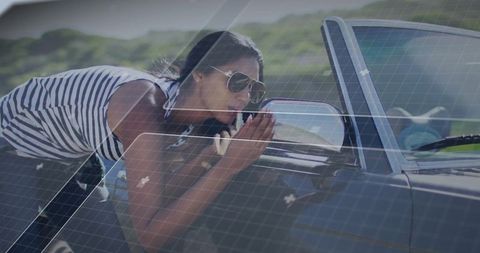 Leaning woman inspecting side mirror of convertible on sunny coastal road wearing aviators