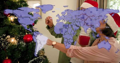 Senior couple decorating christmas tree in santa hats holding red mug and hanging ornaments