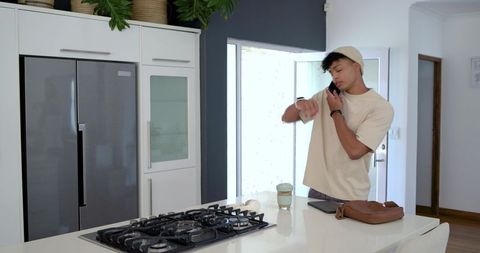 Millennial man checking smartwatch while talking on phone in bright minimalist kitchen