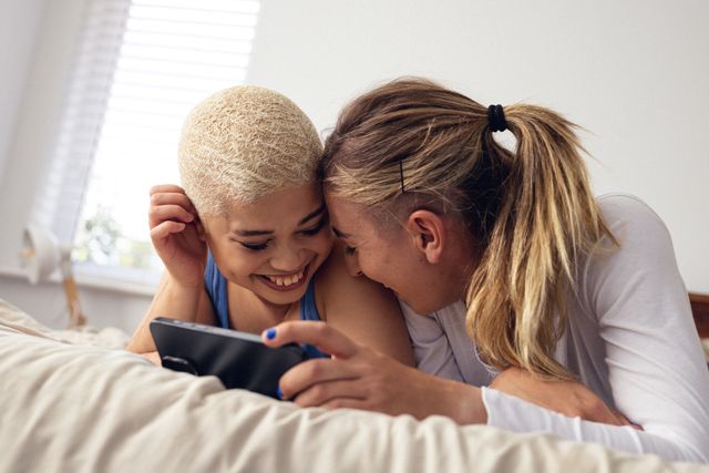 Multiracial lesbian couple lying on bed, laughing while watching something on a cellphone. Perfect for use in articles or advertisements about LGBTQ relationships, technology in daily life, or promoting diversity and inclusion. Can also be used in lifestyle blogs, social media posts, or campaigns celebrating love and togetherness.