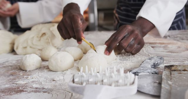 Hands of baker shaping dough balls on floured surface in professional kitchen. Focus on details of baking process and dough preparation, with kitchen tools and flour-covered table adding to the authentic bakery atmosphere. Suitable for illustrating baking techniques, professional cooking environments, artisan bread making, and culinary arts education.