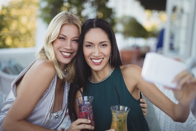 Two women are taking a selfie with a mobile phone at an outdoor restaurant. Both of them are holding colorful drinks and smiling brightly. The image is suitable for use in content related to friendship, leisure activities, summer fun, lifestyle blogs, and social media posts.