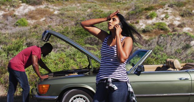 Young woman calling for help while experiencing car trouble on a rural road, with a man inspecting the car's engine. Useful for illustrating car troubles, roadside assistance, mechanical issues, and travel mishaps.