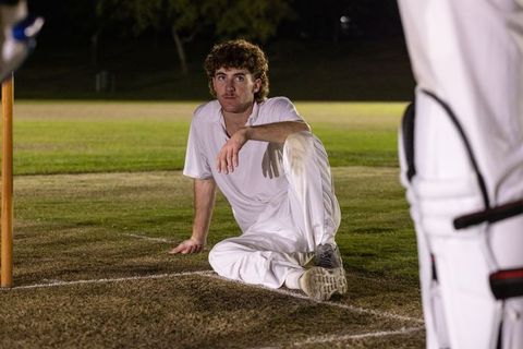 Night cricket player sitting by stumps under floodlights wearing white cricket kit