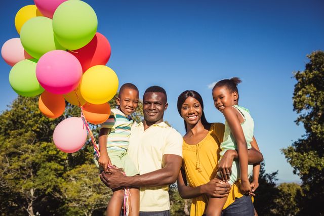 African American family enjoying a sunny day at the park. Parents holding their children and colorful balloons, all smiling and looking happy. Ideal for use in advertisements, family-oriented content, lifestyle blogs, and promotional materials focusing on family bonding, outdoor activities, and happiness.