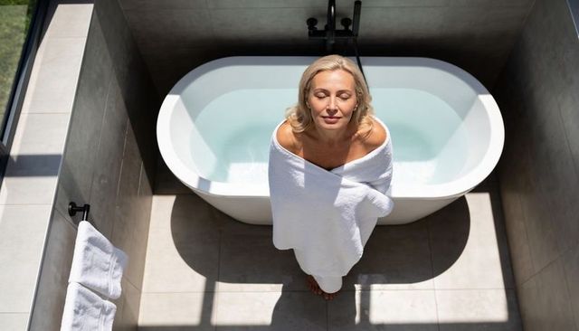Senior woman relaxing wrapped in towel beside freestanding bathtub in sunlit modern bathroom