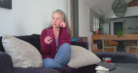 Mature woman sitting on dark sectional sofa checking smartphone looking thoughtful