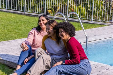 Three friends laughing on wooden pool deck enjoying sunny backyard hangout in casual sweaters