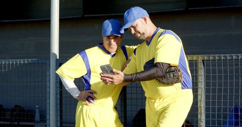 Baseball teammates checking phone in dugout wearing yellow uniforms and blue caps