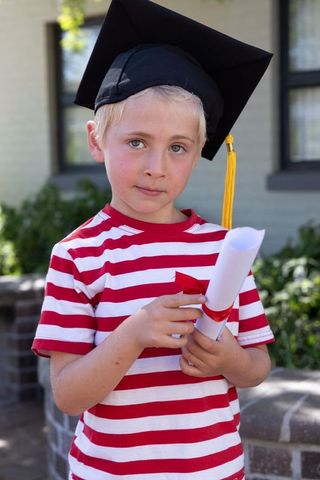 Young child wearing mortarboard and holding diploma roll on porch celebrating graduation