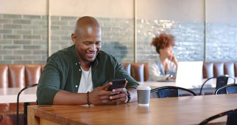 African man smiling while using smartphone at cafe table, woman working on laptop behind