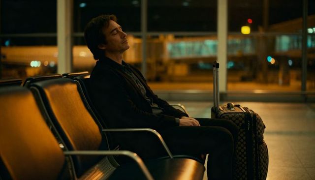 Solo traveler sleeping in late-night airport lounge with suitcase and amber lighting