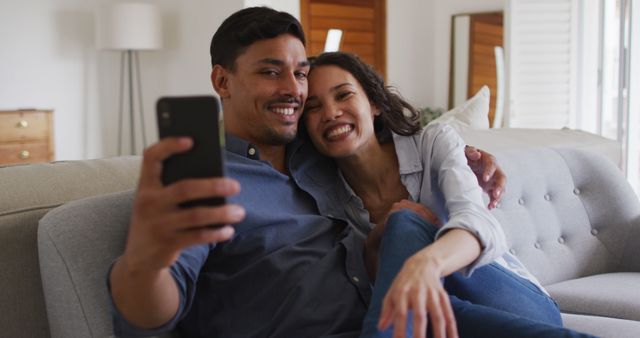 Happy couple sitting on a couch, taking a selfie with a smartphone. They are smiling and appear relaxed and content. Ideal for concepts related to relationships, leisure, happiness, social media, and home life.