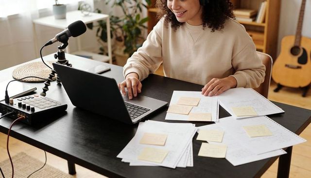 Smiling content creator recording podcast at home desk with microphone, laptop and notes