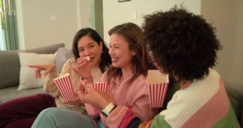 Diverse friends laughing and sharing popcorn on cozy living room couch