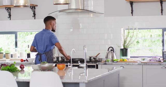 This image showcases a young man cooking in a modern kitchen filled with natural light. The bright, airy setting with an organized countertop suggests a healthy and productive home atmosphere. Ideal for use in advertisements, blogs, and articles about home cooking, healthy eating, kitchen design, or lifestyle content.
