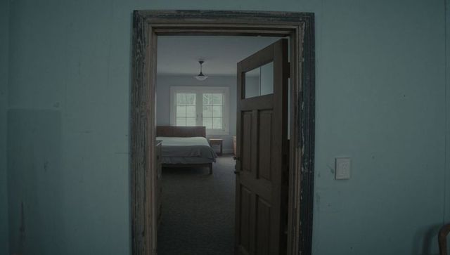 Weathered door framing modest vintage bedroom with made bed and soft neutral daylight mood