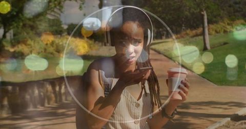 Young woman talking on phone while holding coffee and wearing headphones on campus path