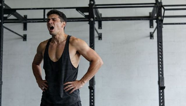Gasping muscular man standing by metal rig wearing black tank recovering from intense workout