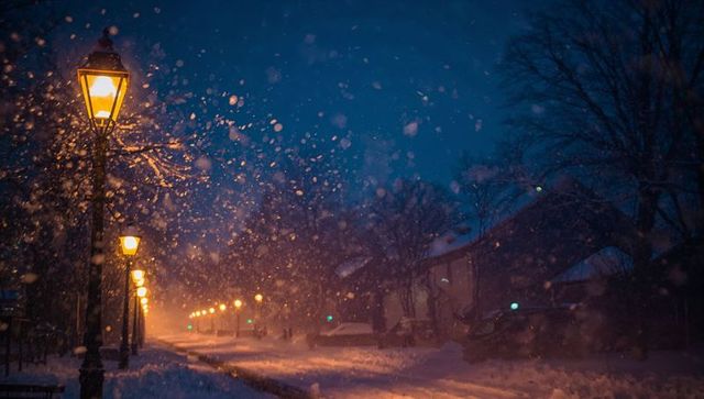 Vintage lampposts glowing amber on snowy neighborhood street at night with falling snow