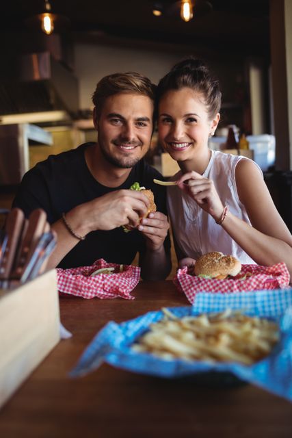 Young couple smiling and enjoying burgers and fries in a cozy bar. Perfect for use in advertisements for restaurants, fast food chains, or lifestyle blogs focusing on relationships and dining experiences.