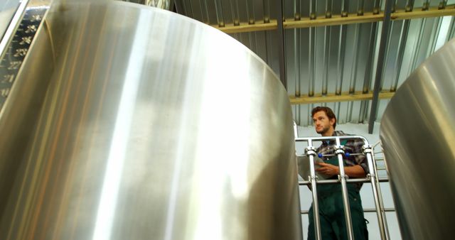 Brewery worker inspecting large stainless steel fermentation tanks in modern beer production facility. Ideal for illustrating brewing processes, manufacturing techniques, industrial environments, or beer and beverage production industries. Suitable for use in articles, blogs, advertisements, and promotional materials related to the brewing industry.