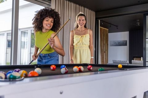 Diverse female friends playing pool at home laughing and enjoying billiards in modern lounge