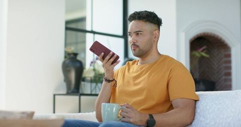 African american man speaking into smartphone while holding coffee mug on couch