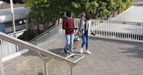 Young diverse travelers walking with carry-on luggage and tablet on sunlit urban bridge