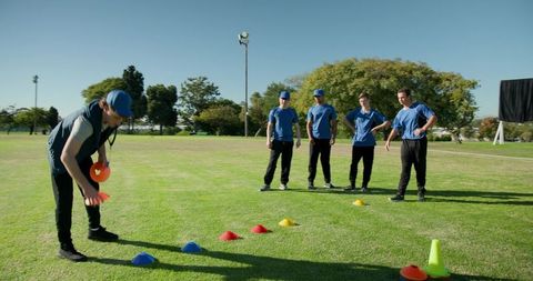 Diverse male team training with cones on grass field observing coach-led teamwork drills