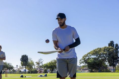 Male athlete practicing cricket batting on sunny park field with bat, ball and helmets