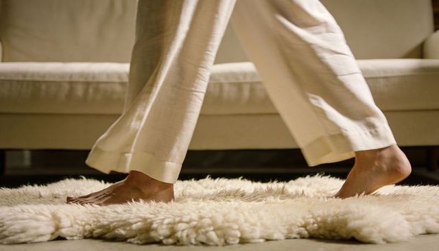 Walking barefoot on plush cream shag rug wearing beige trousers beside sofa
