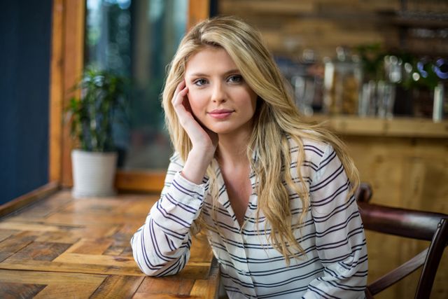 Blonde woman sitting at wooden table in café, leaning on hand, wearing striped shirt. Natural light from window creates a serene atmosphere. Ideal for lifestyle blogs, café promotions, and articles on relaxation or casual fashion.