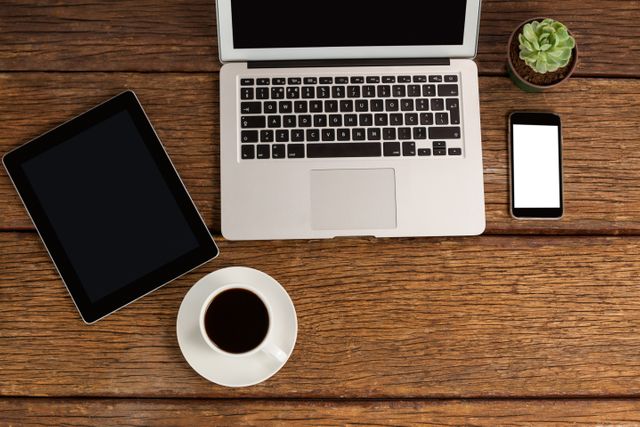 Digital devices including a laptop, tablet, and smartphone are arranged on a wooden table alongside a cup of coffee and a small succulent plant. This setup is ideal for illustrating modern workspaces, remote work, or technology use in everyday life. Perfect for articles, blogs, and advertisements related to productivity, technology, and home office environments.