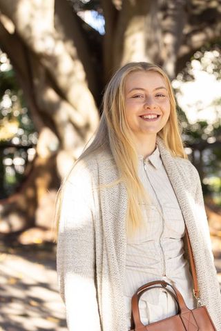 Sunlit woman smiling in park wearing beige cardigan holding brown leather handbag