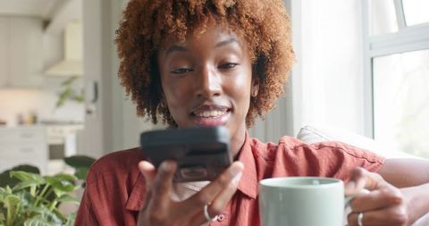 African american woman holding smartphone and coffee mug, smiling on sunlit sofa by window