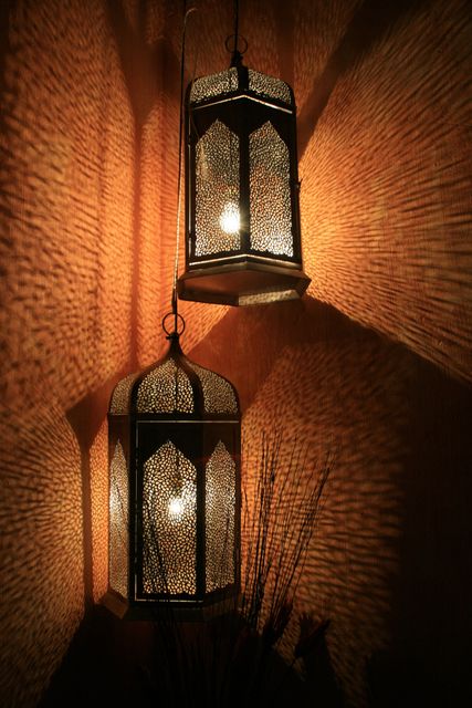 Double Moroccan lanterns hanging from a ceiling emit light, casting intricate shadows on textured walls. Captures warm and inviting ambiance ideal for setups needing an intimate and cozy atmosphere. Perfect for content related to interior design, home decor, oriental themes, exotic culture, or vintage-style indoor lighting solutions.
