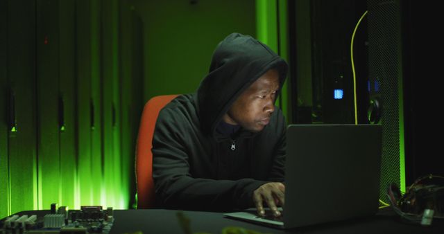 Hacker wearing a hooded jacket sitting in front of a laptop in a dark room with green ambient light. Ideal for illustrating cybersecurity and data privacy threats, hacking activities, and information security breaches.