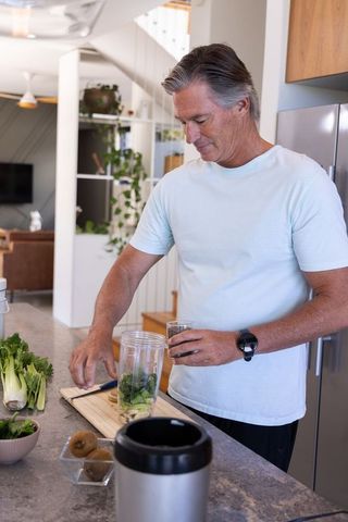 Mature man preparing green smoothie at modern kitchen island with kiwi and leafy greens