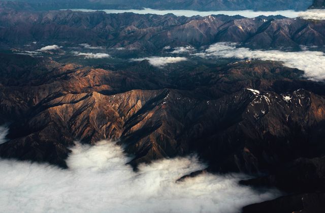 Aerial view capturing the grandeur of mountain ranges enveloped by clouds and mist, showcasing the rugged terrain and serene environment. Ideal for use in travel brochures, nature documentaries, and landscape-themed marketing materials.